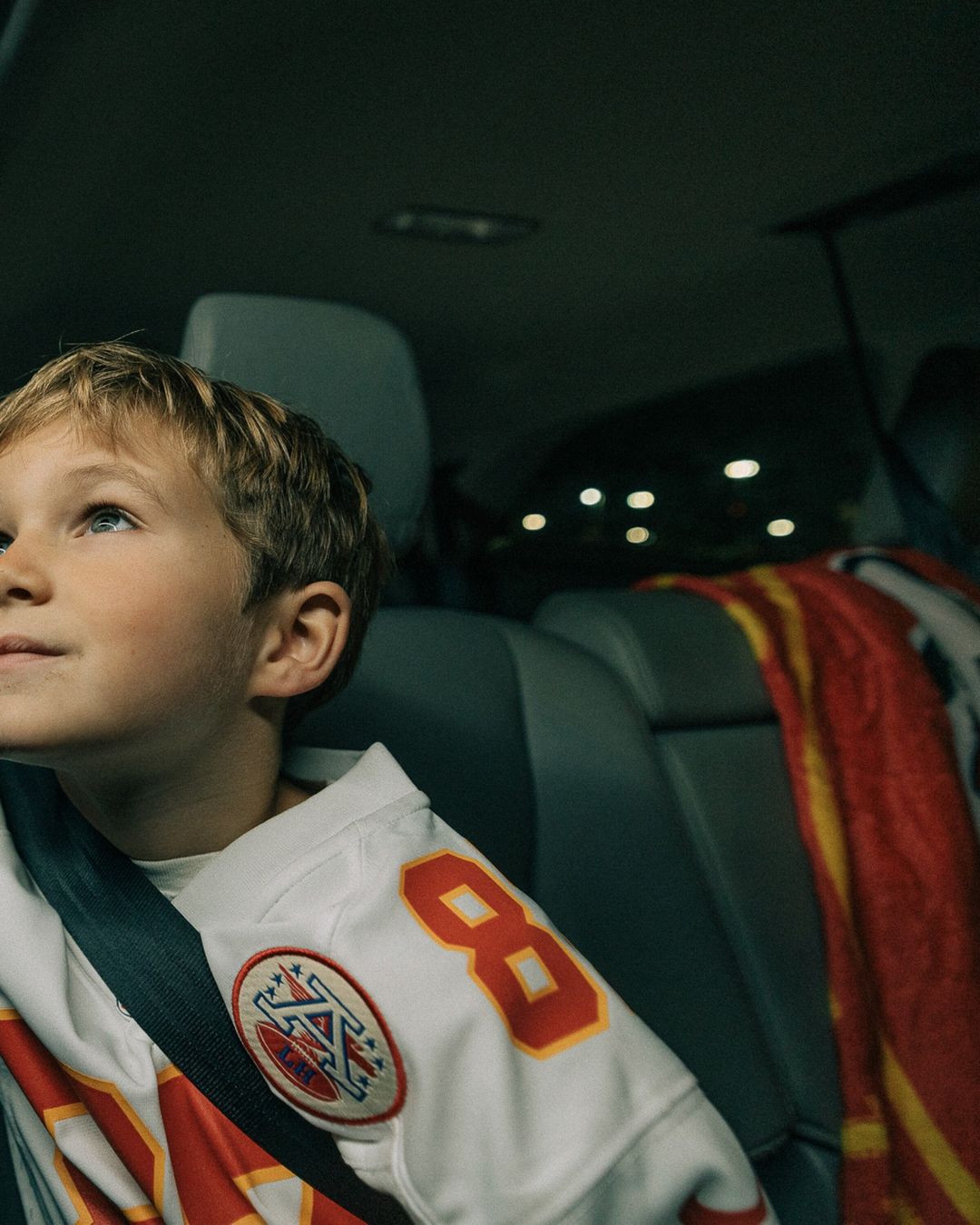 A young boy in a football jersey rides in the back seat of a car, wearing his seatbelt, with a blanket on the seat behind him.