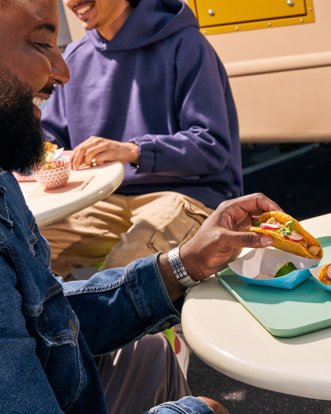 Two people eating tacos outdoors; one person is holding a taco, and the other wears a hoodie, with a food truck visible.