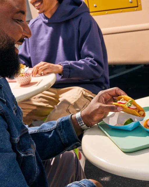 Two people eating tacos outdoors; one person is holding a taco, and the other wears a hoodie, with a food truck visible.