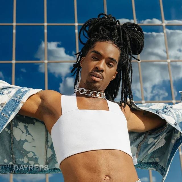 Young African-American man in a white crop top poses outdoors against a glass building with a denim jacket and chain necklace.