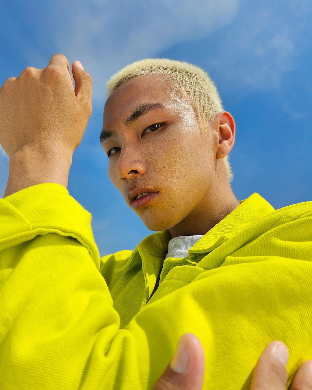 Close-up low angle portrait of an Asian man with bleached blonde hair wearing a bright yellow shirt against a blue sky.