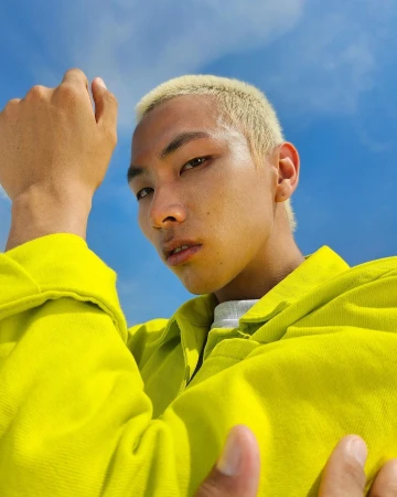 Close-up low angle portrait of an Asian man with bleached blonde hair wearing a bright yellow shirt against a blue sky.