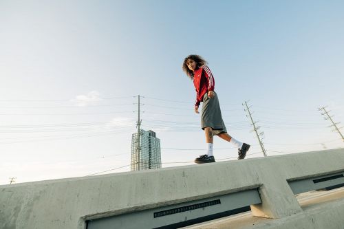 A young adult walks along a concrete structure, wearing a red Adidas track jacket and shorts, with a city building in the background.