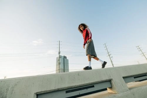A young adult walks along a concrete structure, wearing a red Adidas track jacket and shorts, with a city building in the background.