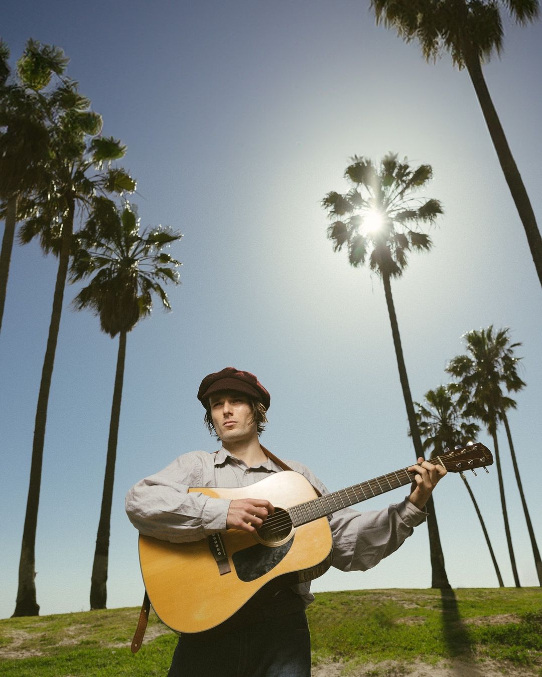 Adult male playing an acoustic guitar outdoors with palm trees and a blue sky, wearing a cap and long-sleeved shirt.