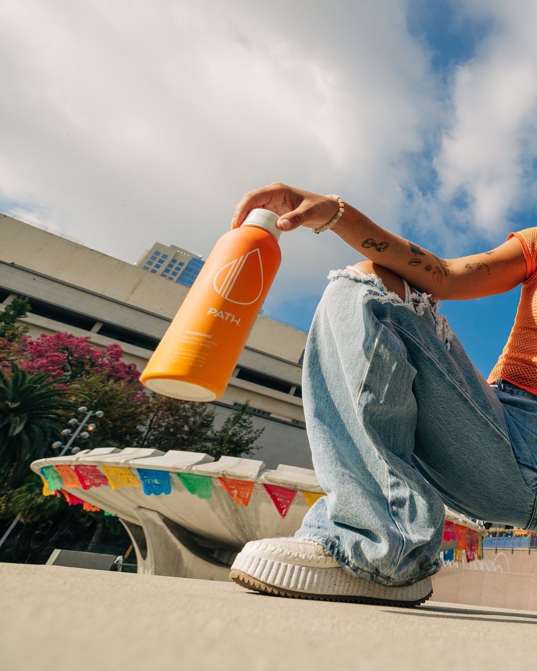 Woman in jeans and white shoes sits in an urban setting with blue sky and a large orange PATH water bottle in hand.