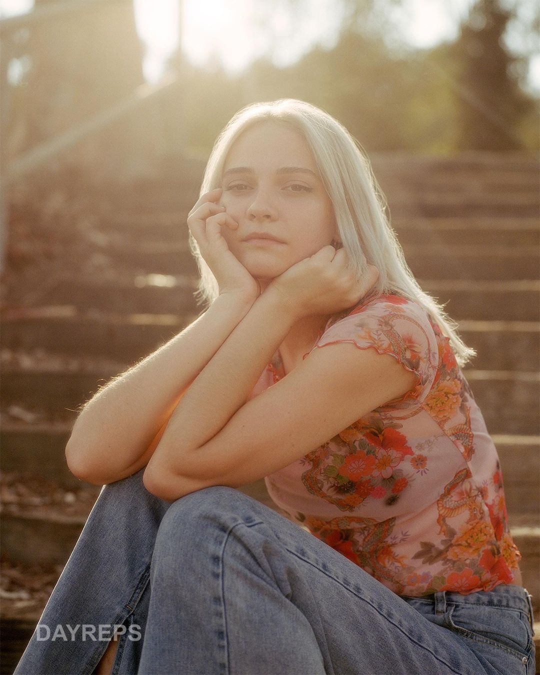 Portrait of a fair-skinned woman with blonde hair sitting on concrete steps wearing a floral top and jeans in warm, natural light.
