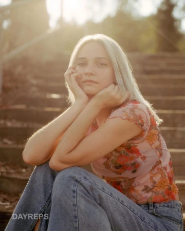 Portrait of a fair-skinned woman with blonde hair sitting on concrete steps wearing a floral top and jeans in warm, natural light.