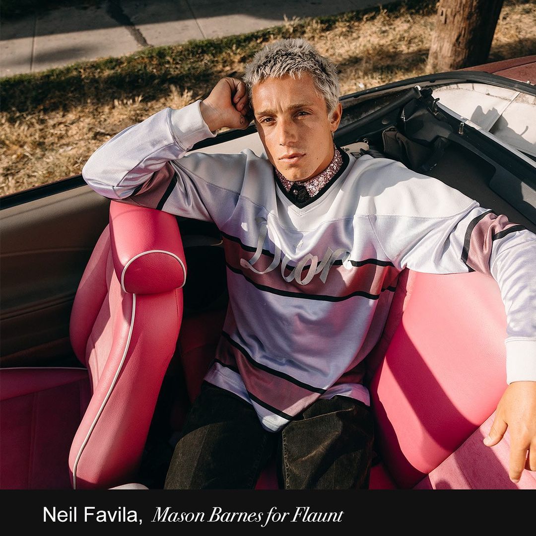 Young adult poses in the pink leather interior of a vintage convertible car on a sunny street with houses in the background.