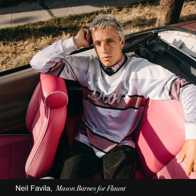 Young adult poses in the pink leather interior of a vintage convertible car on a sunny street with houses in the background.
