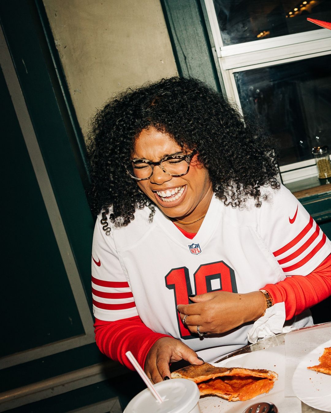 Smiling woman in sports jersey eating pizza indoors. She wears glasses and a gold necklace, and has dark curly hair.