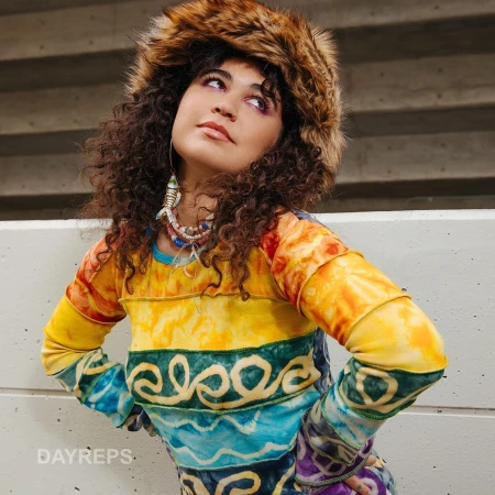 Woman wearing a fuzzy faux fur hat and colorful patterned top posing outdoors against a concrete wall