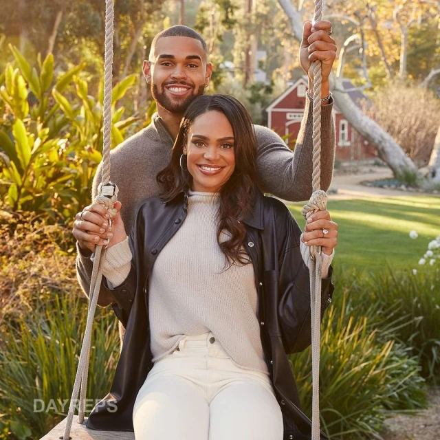Smiling couple on swing in sunlit outdoor setting. Brunette woman in jacket; man behind. Green grass, trees, red house in background.
