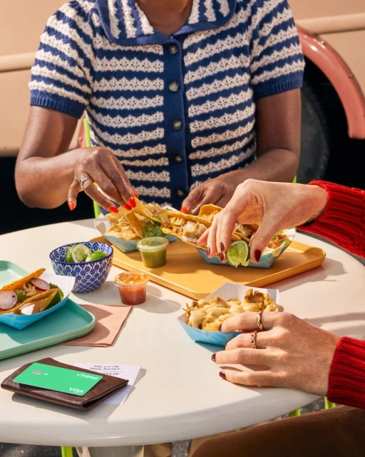 Two people share tacos and appetizers at a table outside, with a wallet and restaurant bill in view, bathed in natural light.