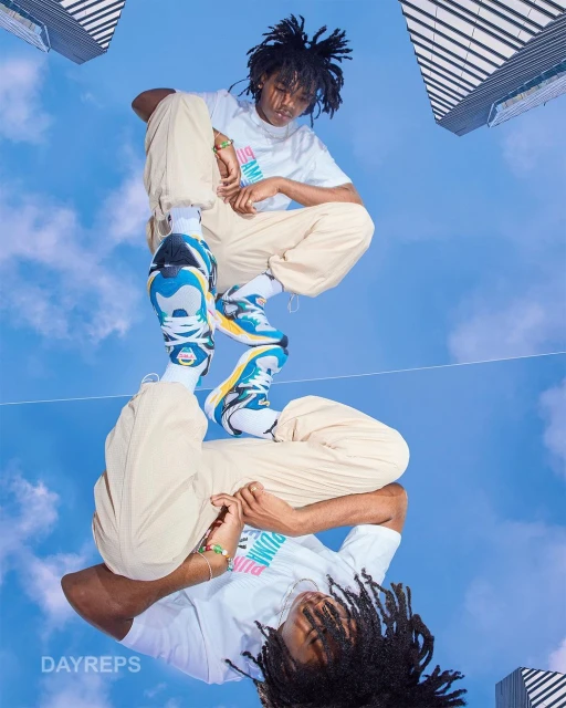 Young man in beige pants and a white t-shirt posing with sneakers, reflecting the sky and modern buildings in the background.