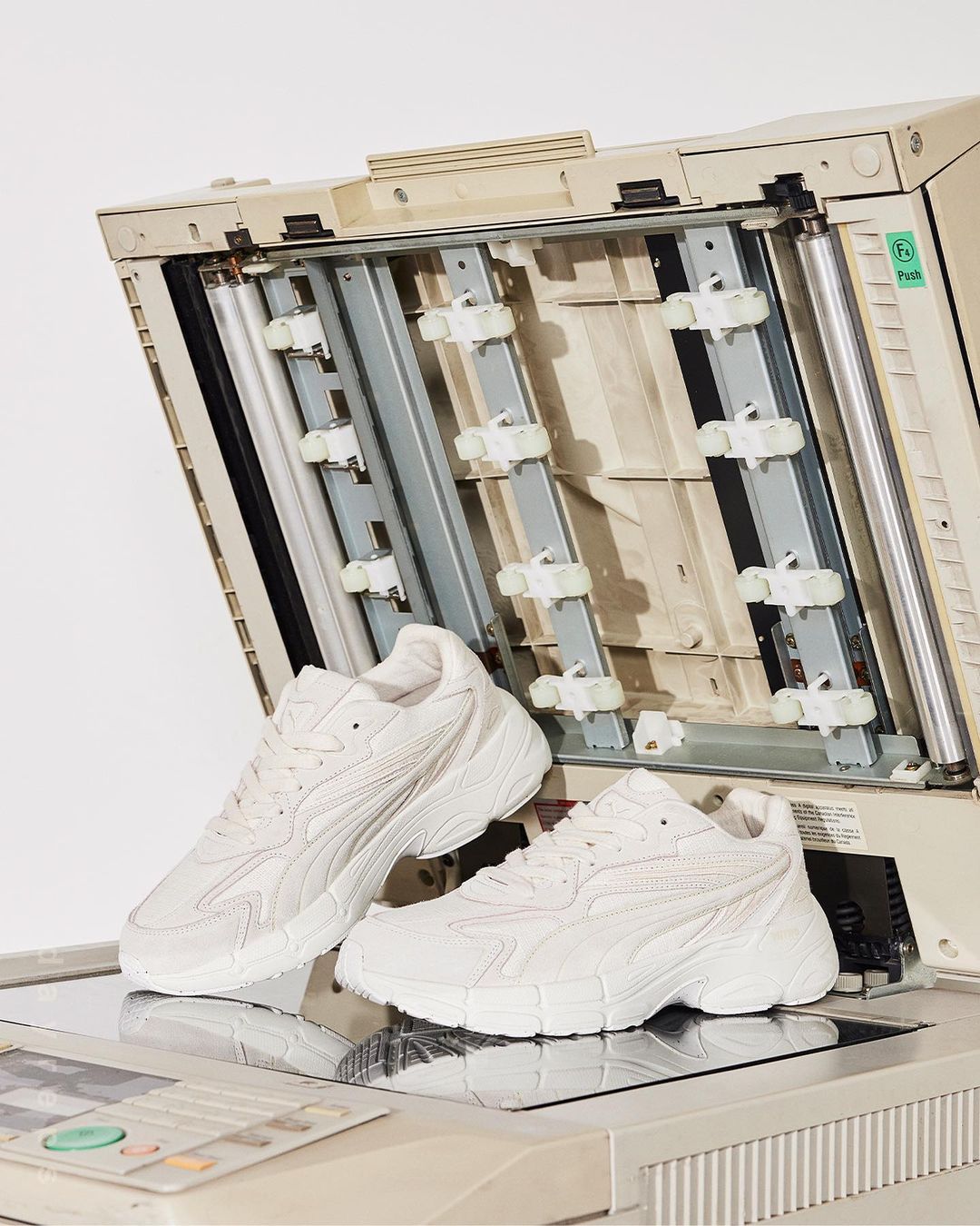Two white sneakers are displayed on top of an open office copier in a photo studio setting, with the copier's internal mechanism visible.