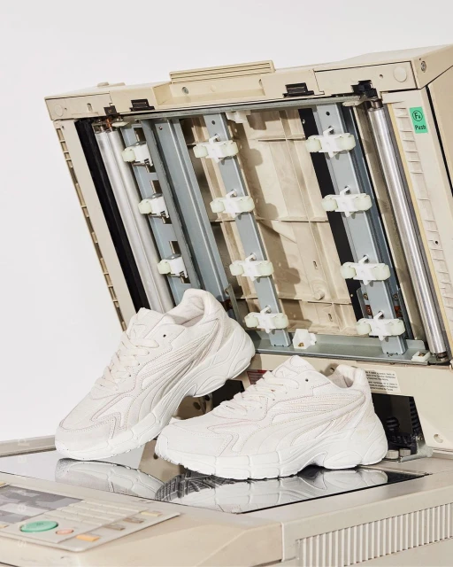 Two white sneakers are displayed on top of an open office copier in a photo studio setting, with the copier's internal mechanism visible.