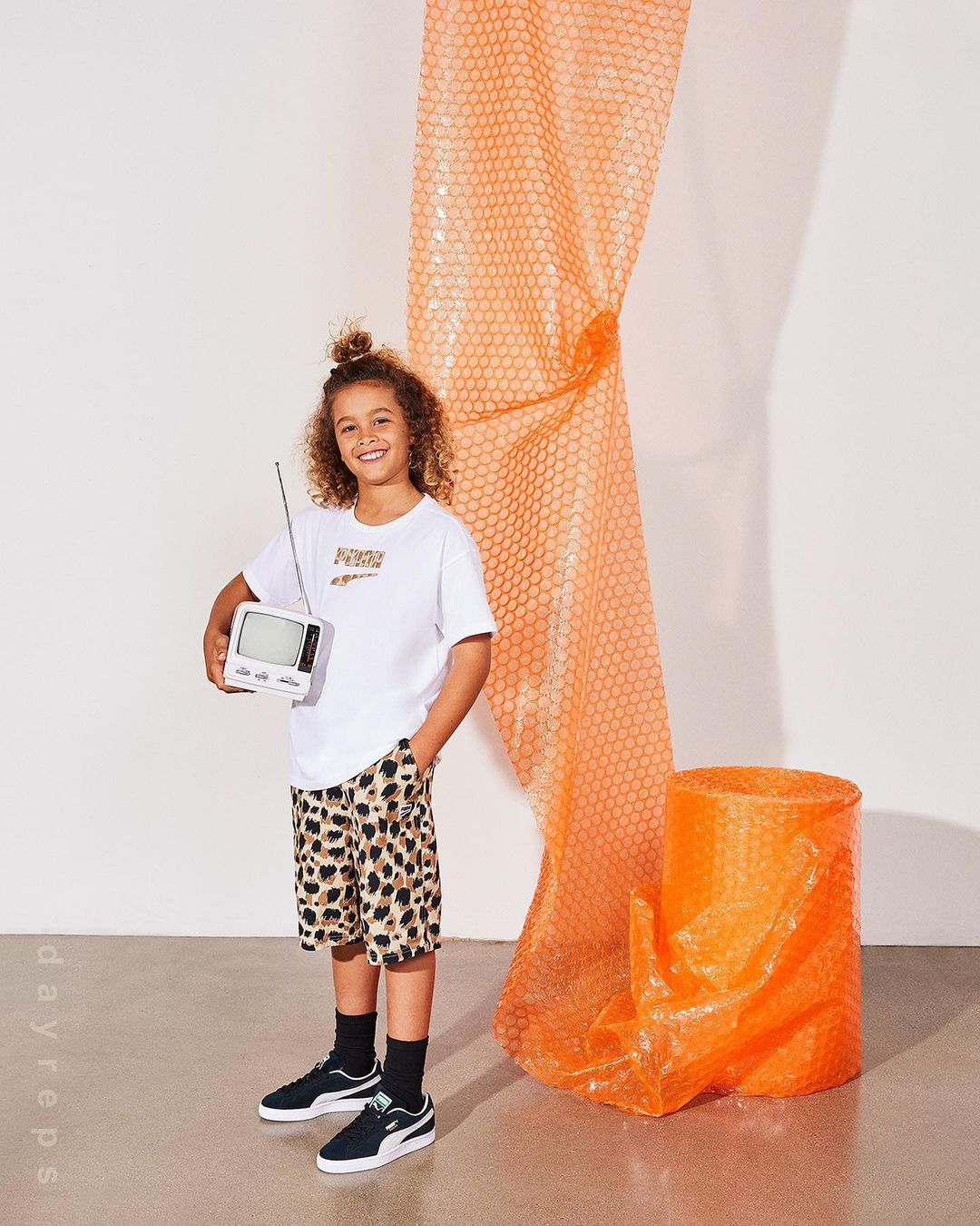 Young boy in leopard-print shorts holds a retro TV against a white background with an orange bubble-wrap roll and cylinder prop.