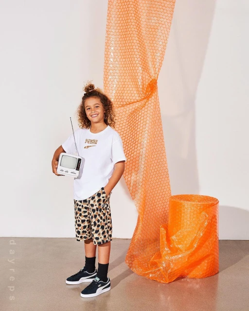 Young boy in leopard-print shorts holds a retro TV against a white background with an orange bubble-wrap roll and cylinder prop.