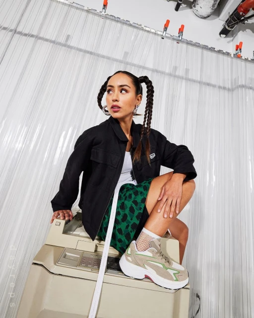 A stylish young woman with braided pigtails and a black jacket poses while sitting on a vintage copy machine in a studio setting.