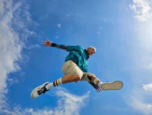 Adult male jumping in the air with arms outstretched, wearing a turquoise jacket, tan shorts, and sneakers against a blue sky backdrop.