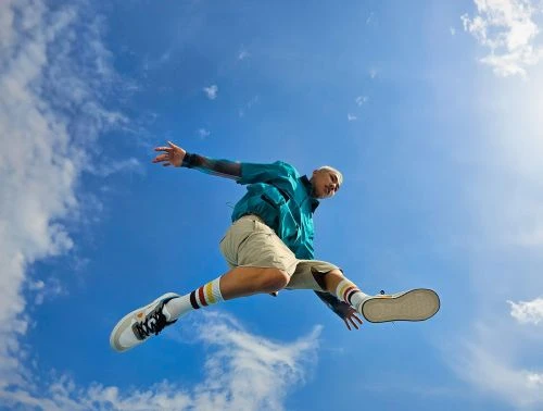 Adult male jumping in the air with arms outstretched, wearing a turquoise jacket, tan shorts, and sneakers against a blue sky backdrop.