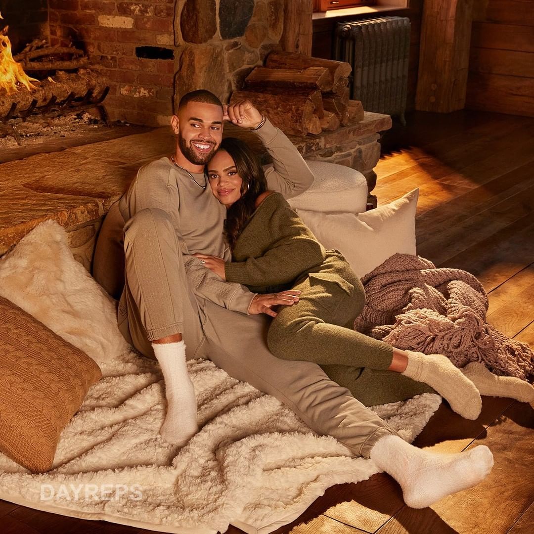 A smiling couple, a man and a woman, are sitting together on a blanket in front of a fireplace, wearing matching loungewear.