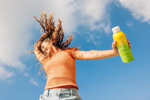 Woman smiles broadly with arms outstretched holding a yellow and green drink bottle against a blue sky with puffy white clouds.