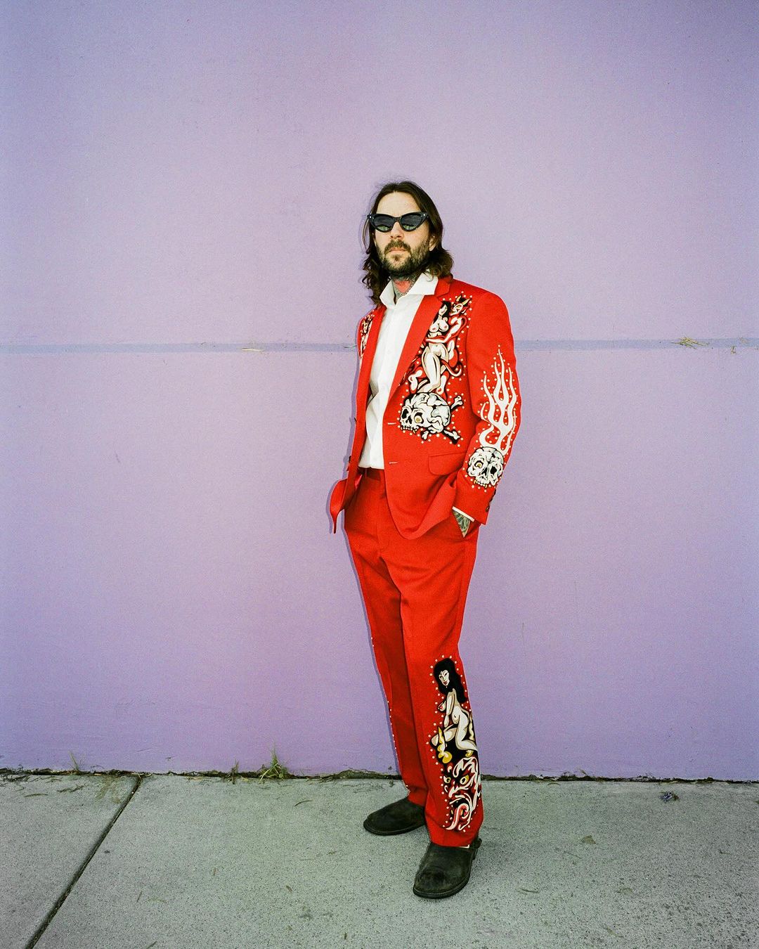 Man in a red suit with custom art posing against a purple wall on a city street with sidewalk. He wears black sunglasses.