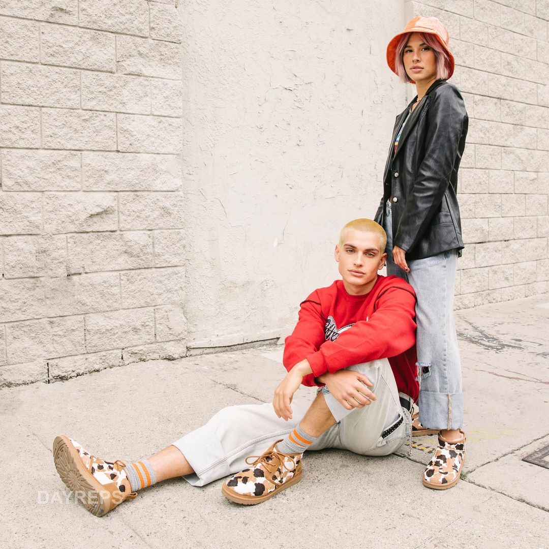 Two people posing outdoors in casual fashion. A male sits on the ground while a female stands behind him near a concrete wall.