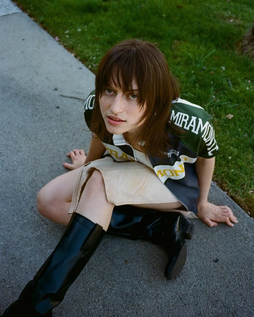 Young adult woman poses outdoors wearing sport-style top, mini skirt, and knee-high black boots against a concrete and grassy background.