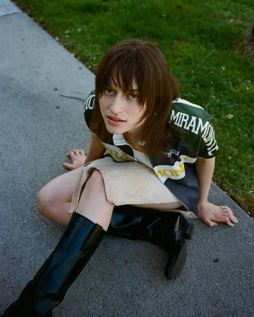 Young adult woman poses outdoors wearing sport-style top, mini skirt, and knee-high black boots against a concrete and grassy background.