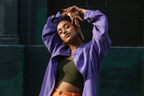 Portrait of a woman in a purple jacket with her hands above her head, standing outdoors against a green wall on a sunny day.