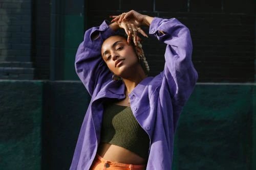 Portrait of a woman in a purple jacket with her hands above her head, standing outdoors against a green wall on a sunny day.