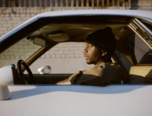African-American man driving classic white car, wearing a black beanie against a blurred brick wall background, golden hour lighting.