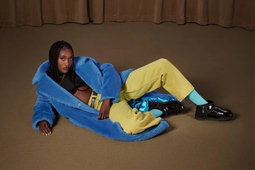 A woman with dark skin and braided hair is lying down wearing a blue furry coat and yellow pants with black boots in a studio setting.