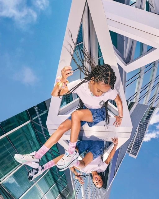 Young woman poses with a reflection in white sneakers, denim shorts, and a white t-shirt in an urban setting with high-rise buildings.