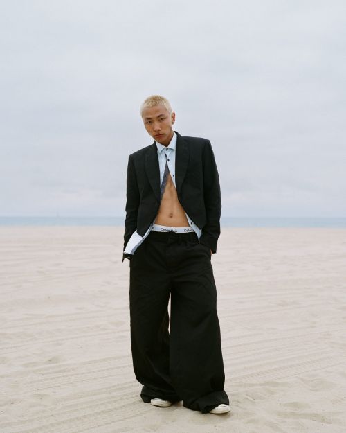 A young adult poses on a beach, wearing a black suit jacket and wide-legged pants, with a visible white undershirt and tie.