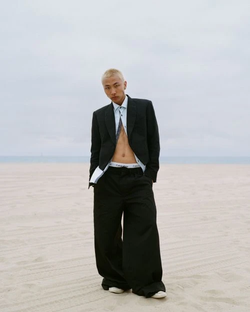 A young adult poses on a beach, wearing a black suit jacket and wide-legged pants, with a visible white undershirt and tie.