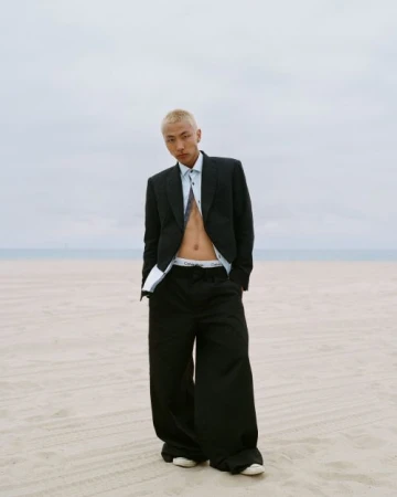 A young adult poses on a beach, wearing a black suit jacket and wide-legged pants, with a visible white undershirt and tie.