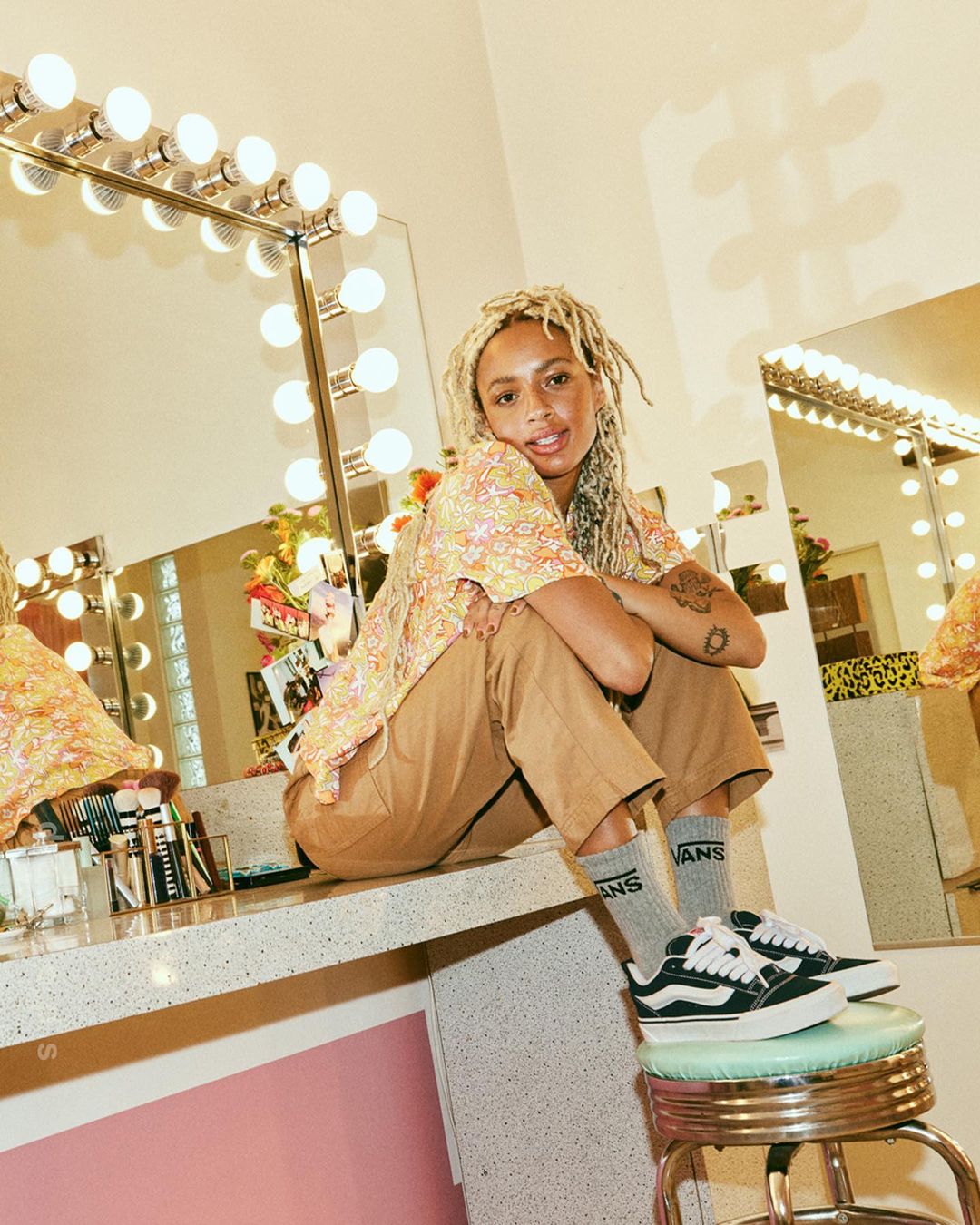Woman with blonde dreadlocks poses in a floral top and brown pants, sitting on a stool near a vanity mirror in an indoor setting.