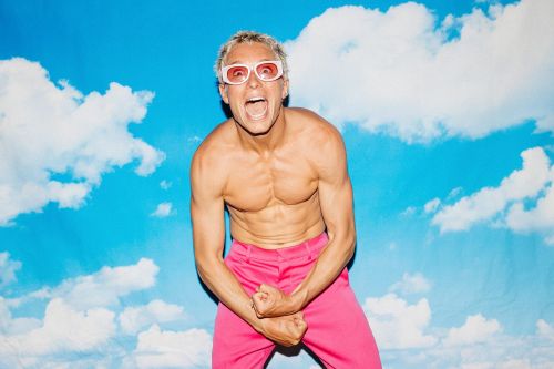Shirtless man with blonde hair and pink glasses shouting with flexed biceps in pink pants against a blue sky and cloud backdrop.