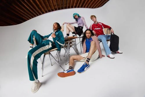 Group of five diverse young adult models sitting on metal chairs wearing Puma branded athleisure clothing in a light studio setting.