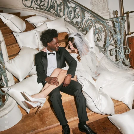 Wedding couple in formalwear sitting on stairs covered with pillows. Bride wears white gown and veil, groom wears black tuxedo.