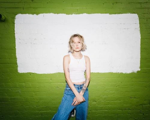 A blonde adult woman with tattoos poses confidently against a vibrant green brick wall with a white painted rectangle detail.