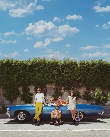 Five people with a vintage blue convertible car in front of a lush green hedge under a sunny sky with fluffy white clouds.