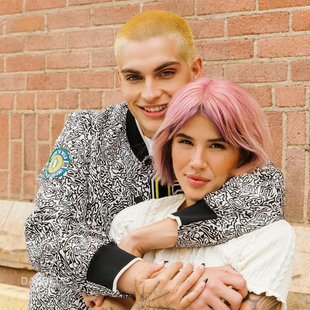 A couple embraces in front of a brick wall: a man with light hair in a patterned jacket and a woman with pink hair in a white sweater.