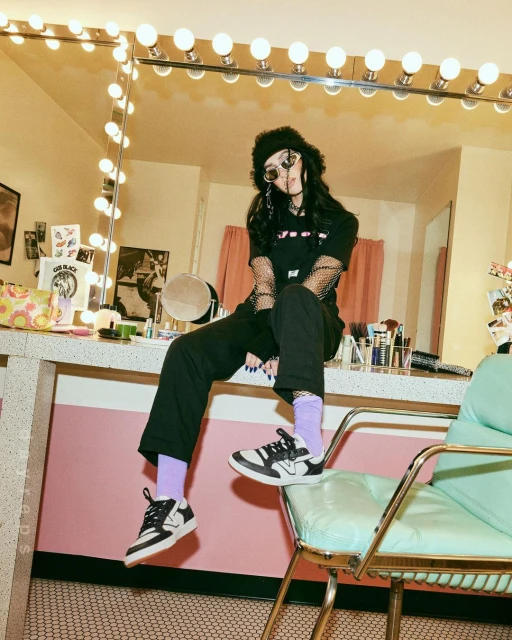 Teen in black streetwear poses on a makeup station chair, framed by a mirror with illuminated bulbs against a pink and yellow backdrop.