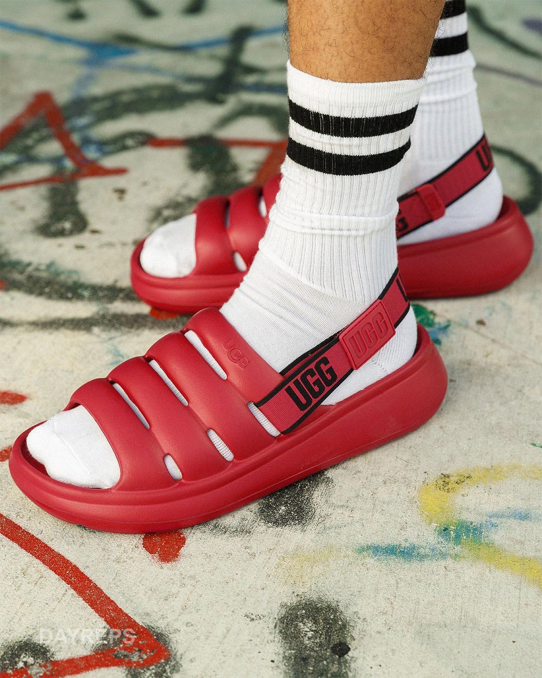 Close-up of a person wearing white socks with black stripes and red UGG sandals, posing on a concrete surface with graffiti.
