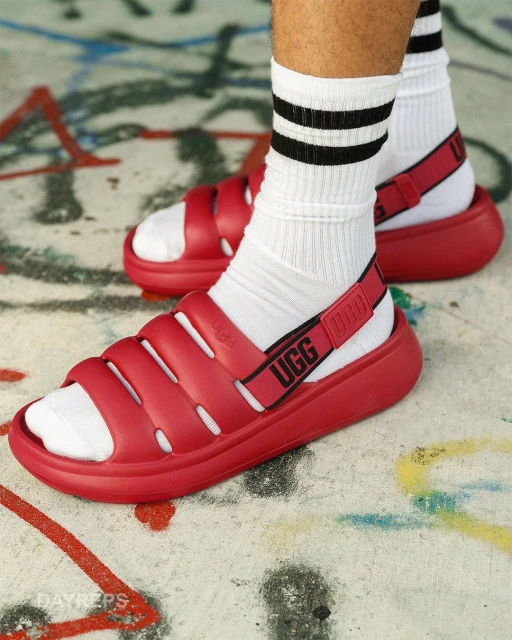 Close-up of a person wearing white socks with black stripes and red UGG sandals, posing on a concrete surface with graffiti.